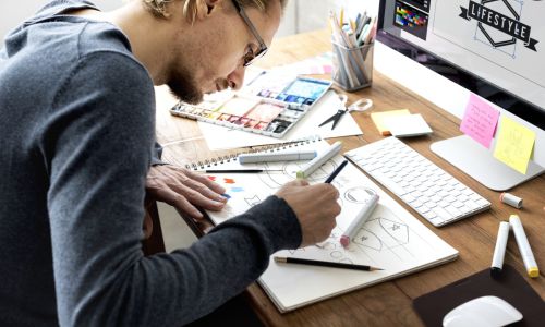 Man sitting at a desk with a computer sketching on paper.